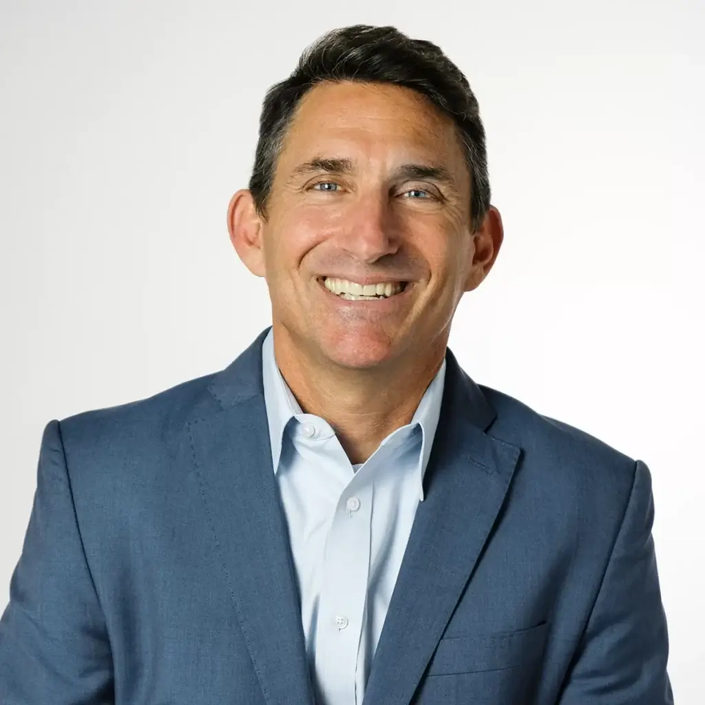 Mark Freid is smiling and wearing a blue sport coat and a light blue shirt while sitting in front of a light-colored background.