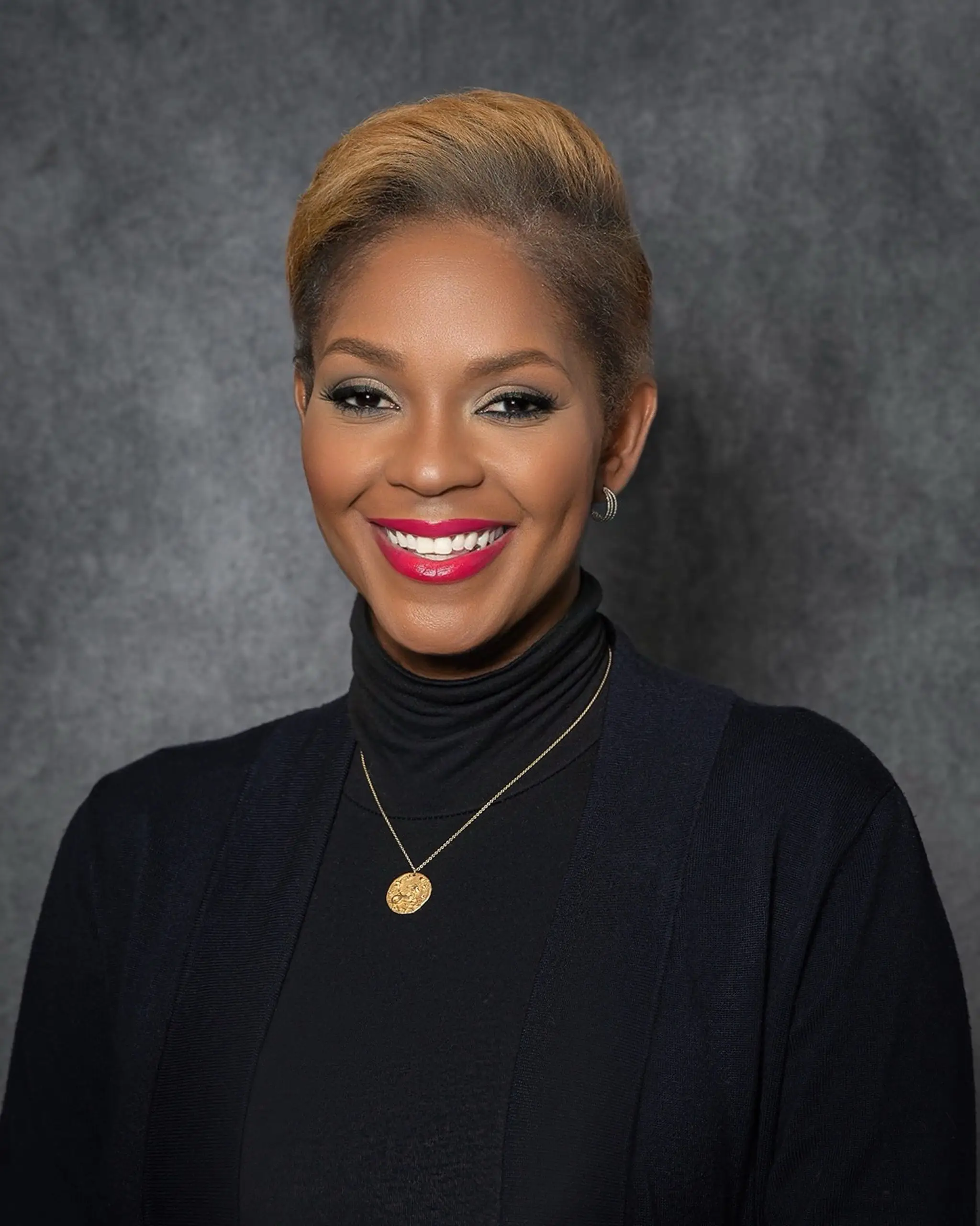 Ericka Dunlap is smiling and wearing a black turtleneck top and a golden chain with a pendant while sitting against a dark-colored background