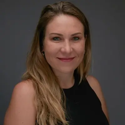 Aleksandra Cuprys is smiling and has long blonde hair while wearing a dark-colored sleeveless top and is sitting against a dark-colored background