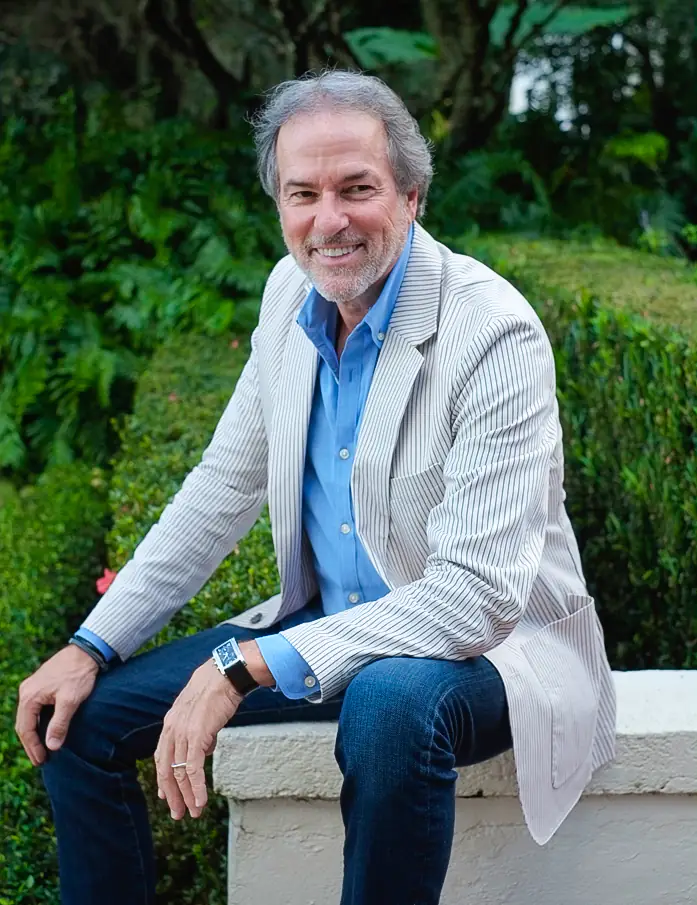 A smiling man with a beard, wearing a blue shirt and a striped blazer, sits on a stone wall in a garden at a Central Florida nonprofit.