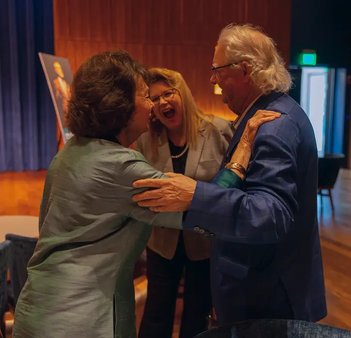 Three guests greeting one another warmly beside a stage.