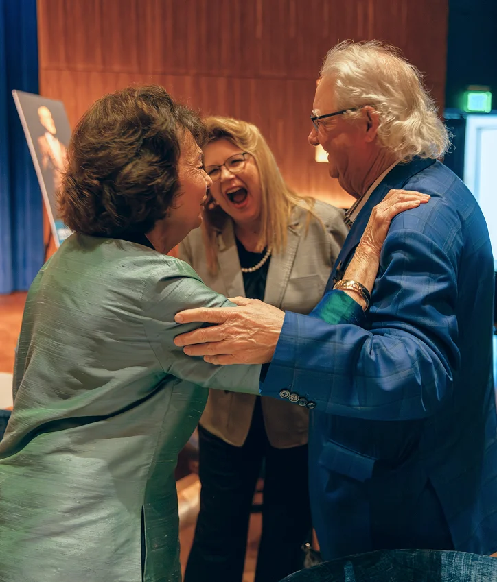 Three guests greeting one another warmly beside a stage.