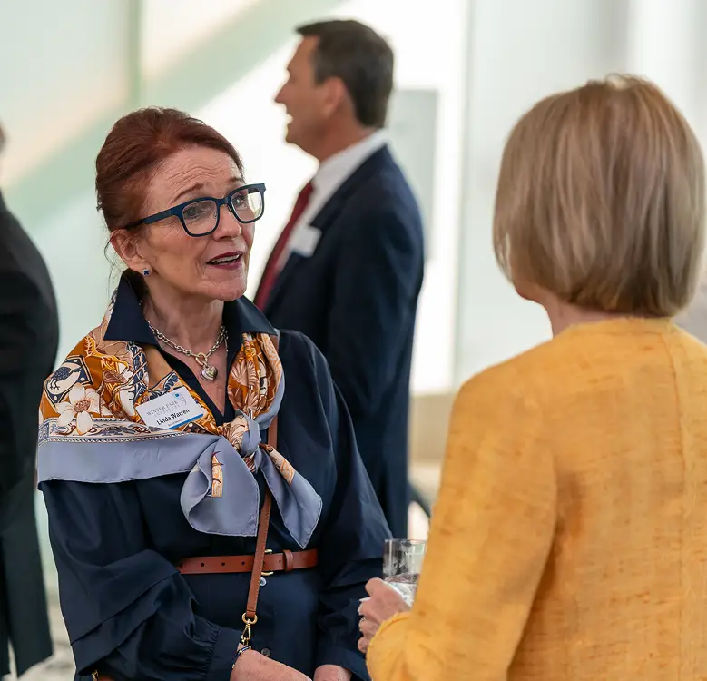 Two women talking at an indoor reception, one wearing a patterned scarf and the other a yellow jacket.