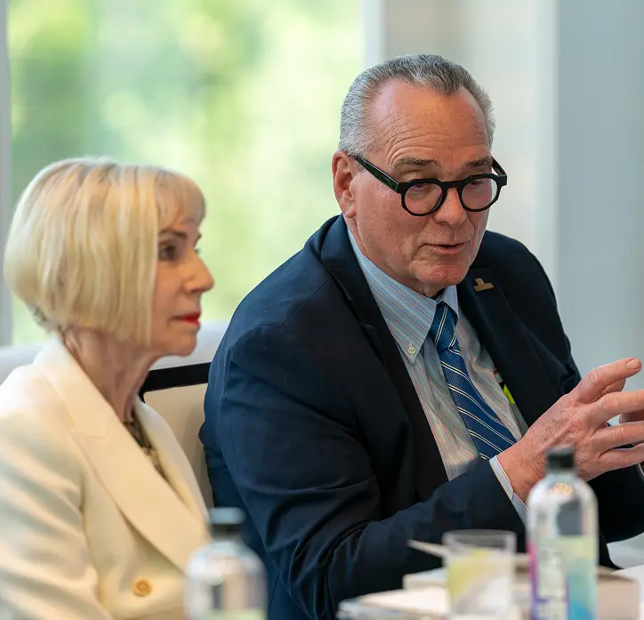 A man in a suit gesturing during a seated discussion beside a woman in a white jacket.