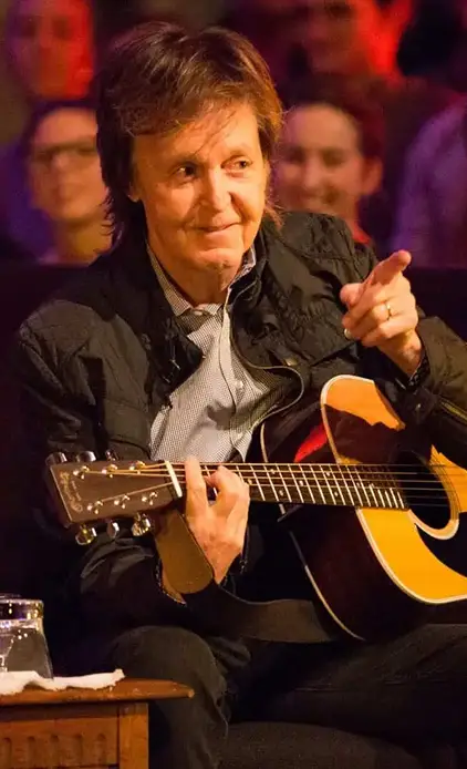 Paul McCartney holding an acoustic guitar and pointing toward someone off camera.