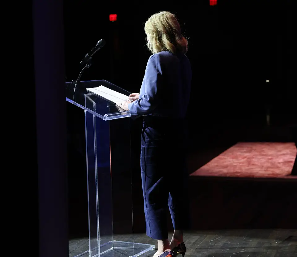 A woman reading from notes at a clear lectern on a darkened stage.