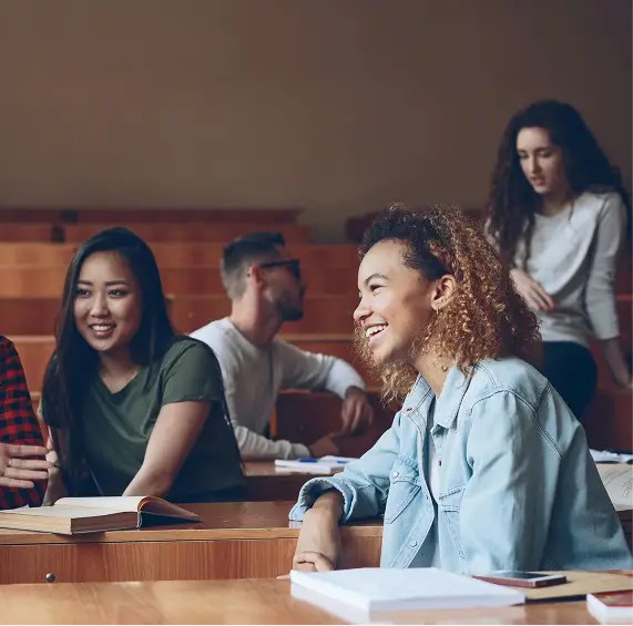 Students smiling and talking in a lecture hall with open books on their desks.