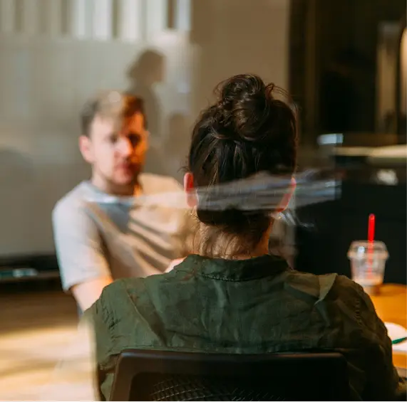 Two people sitting across a table in conversation, viewed through a window.