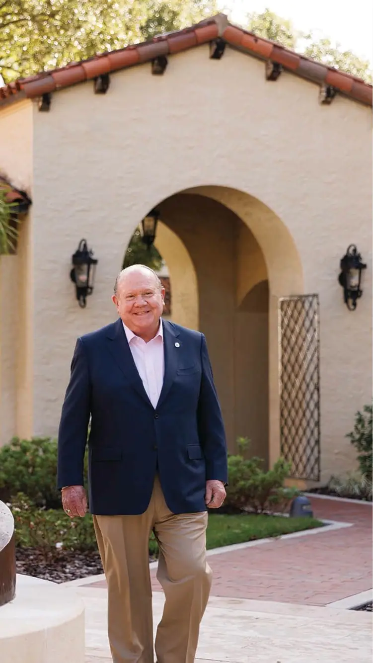 A smiling man in a navy blue blazer and tan trousers standing in front of a Central Florida nonprofit building with an arched entryway.
