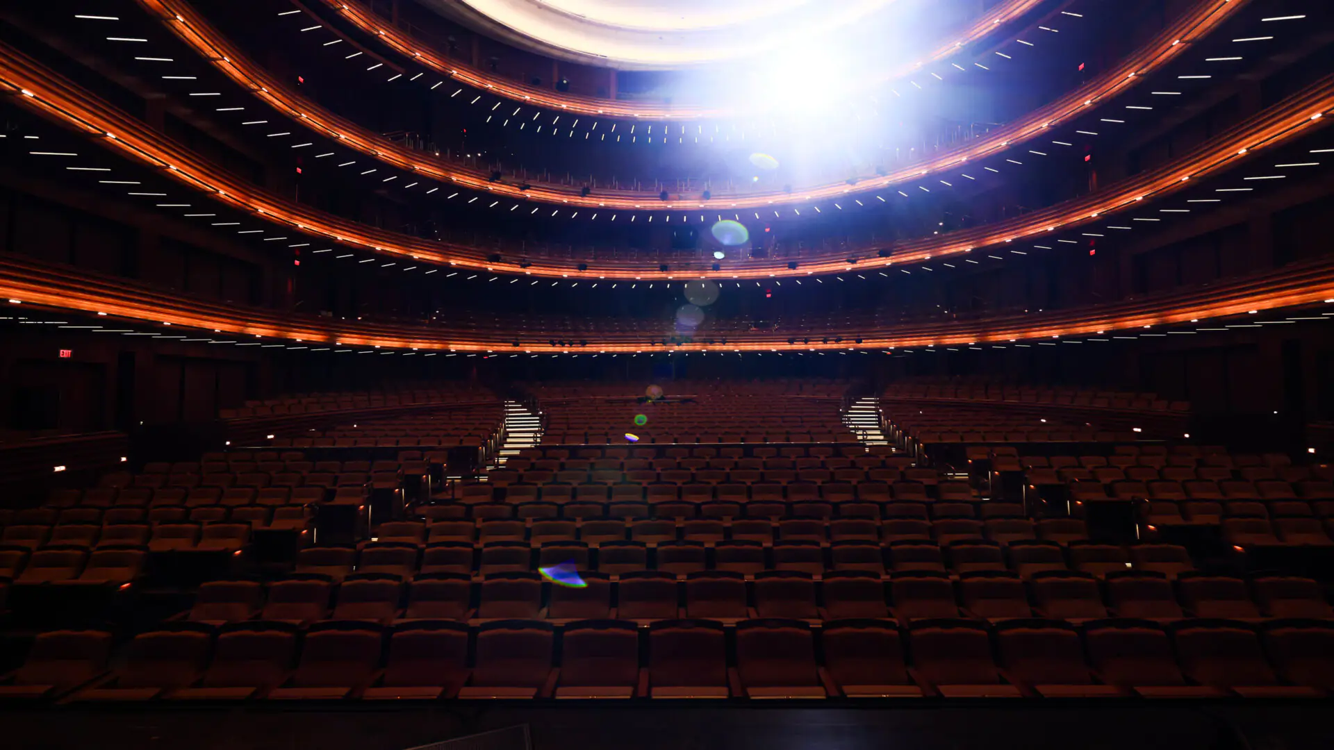 Empty tiered seating inside a large theater with bright lights shining from the stage.