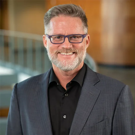 Mark Allen Hayes, a man with a graying beard and blue-framed glasses, wearing a dark blazer and black shirt.