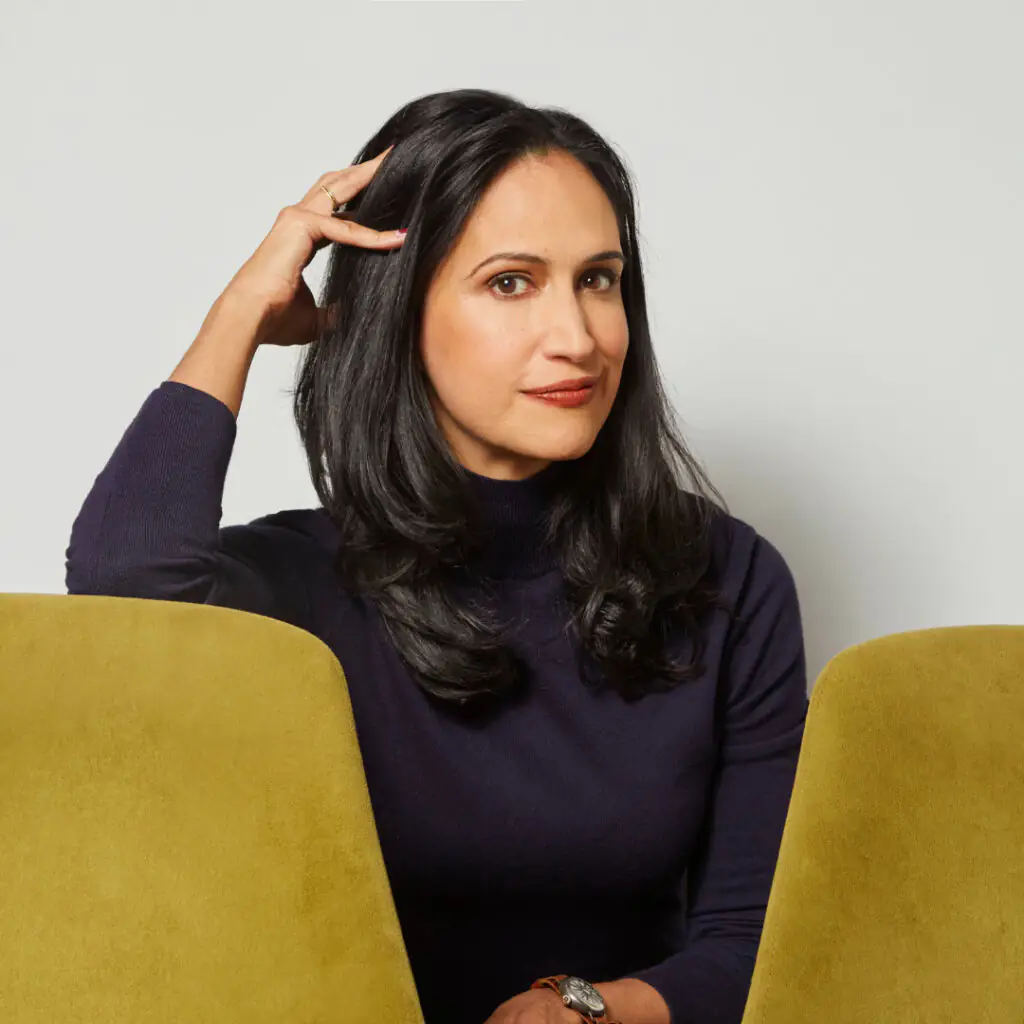 Priya Parker rests her hand on her head while sitting between two yellow chairs, wearing a navy turtleneck sweater.